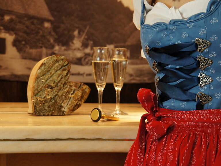 Eine Dame in blauem Dirndl steht vor einem Tisch mit zwei Sektgläsern