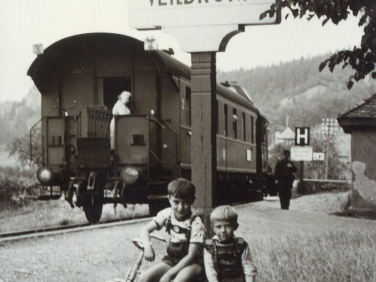 Zwei Kinder stehen vor einem historischen Zug und dem Ortschild von Veilbronn