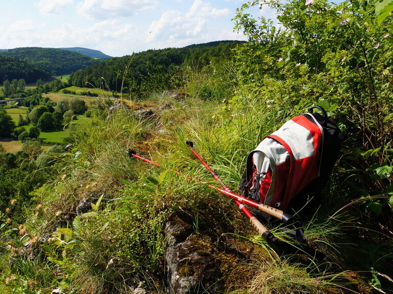 Ein Rucksack sowie zwei Wanderstöcke auf dem Boden, im Hintergrund Ausblick auf die grüne Landschaft der Fränkischen Schweiz